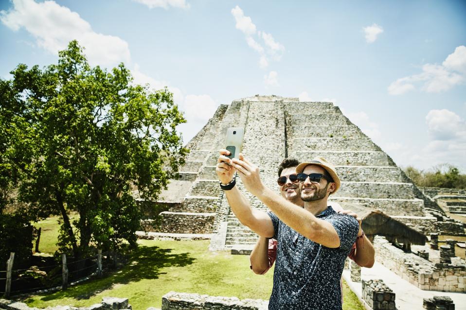 Smiling gay couple taking selfie with smartphone while exploring Mayapan ruins during vacation