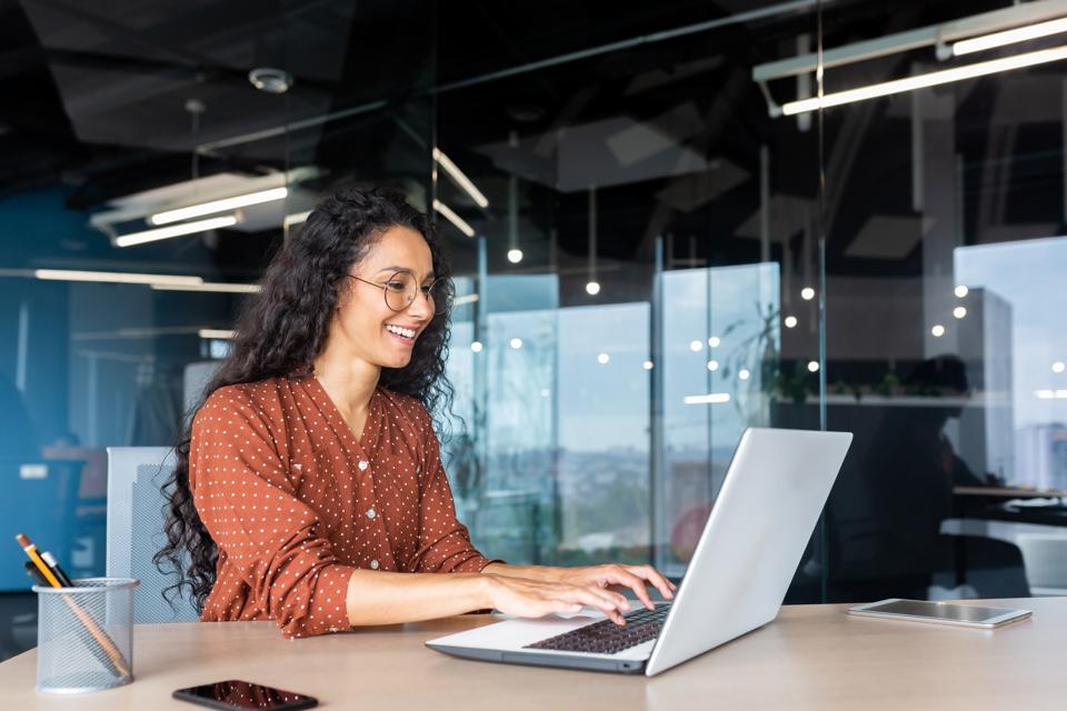 Happy and smiling Hispanic businesswoman typing on laptop in office