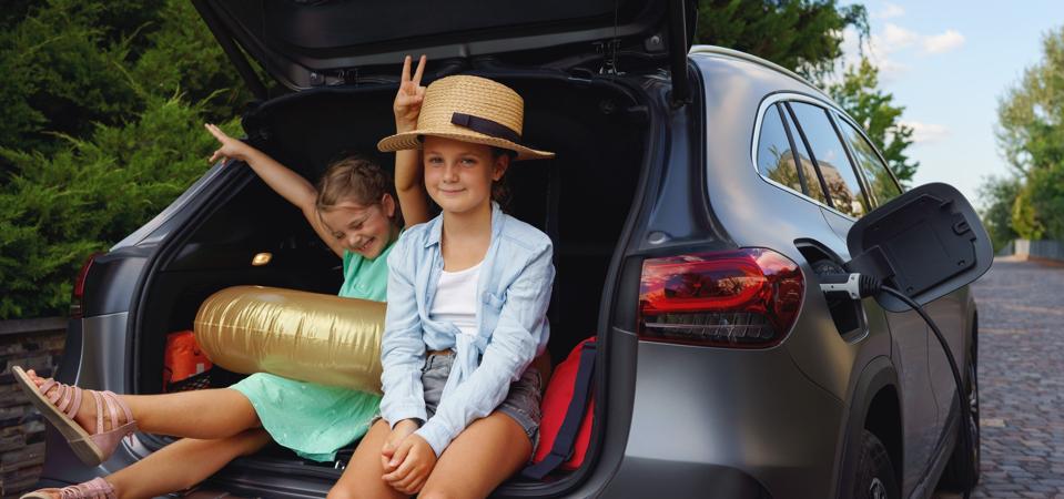Two young girls sitting in the open trunk of a car while it charges during a road trip.