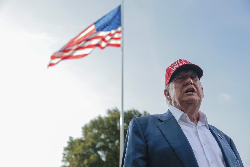 President Trump wearing a red cap speaks in front of a US flag