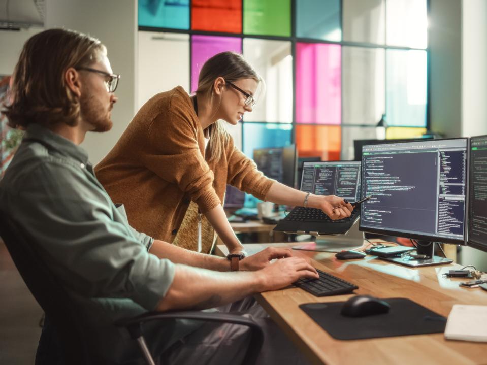 Male Programmer Shows Progress to Female Start-Up CEO on Desktop Computer in Creative Office Space. Caucasian Man and Woman Discussing New Features For Artificial Inteligence Software.