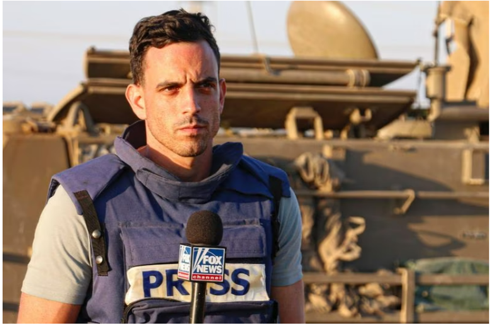 Trey Yingst, a Fox News foreign correspondent, wearing a blue press vest and holding a Fox News microphone while reporting from a military zone with armored vehicles in the background.
