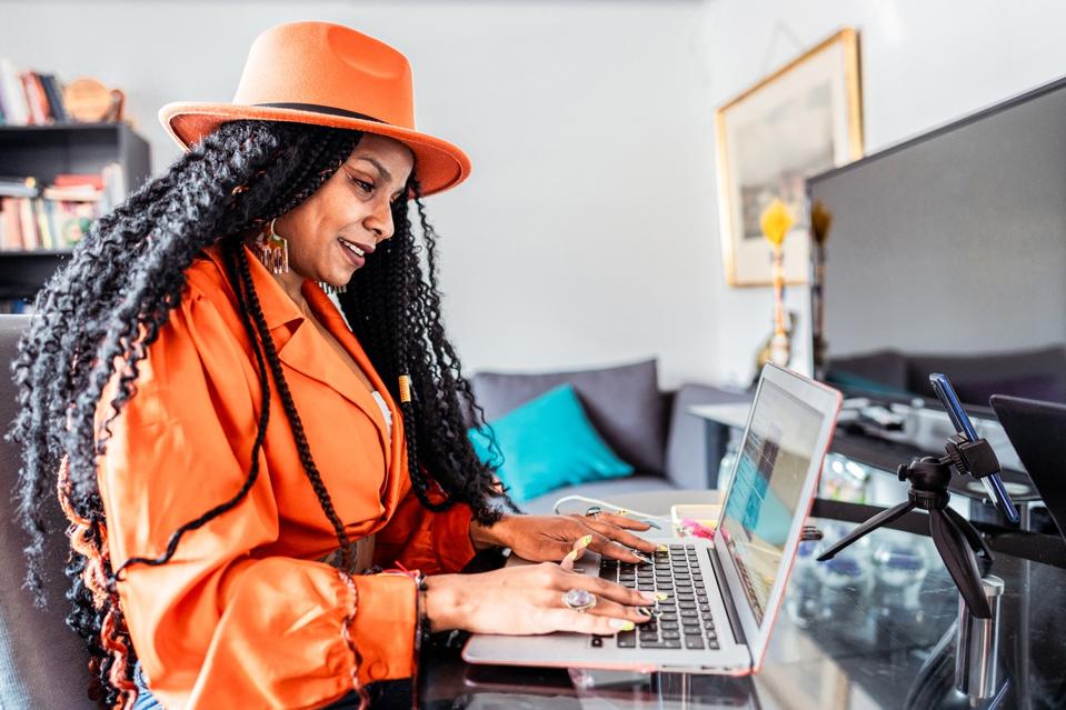 Black woman in orange hat working remotely on a laptop in a modern, colorful home office