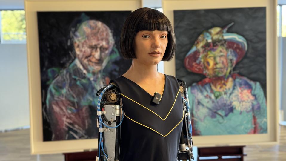 A lifelike humanoid robot in front of a portrait of King Charles and one of Queen Elizabeth II