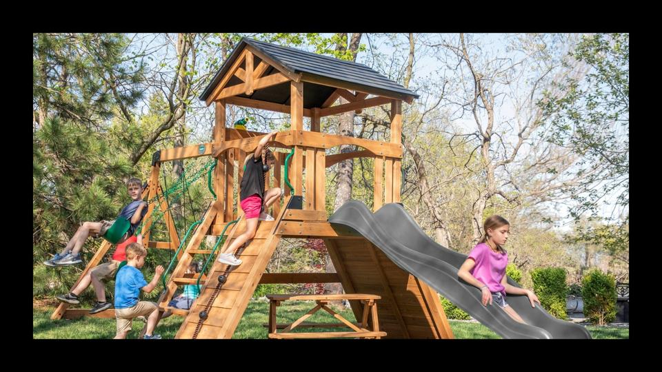 Kids playing on a wooden swing set outside with trees around.