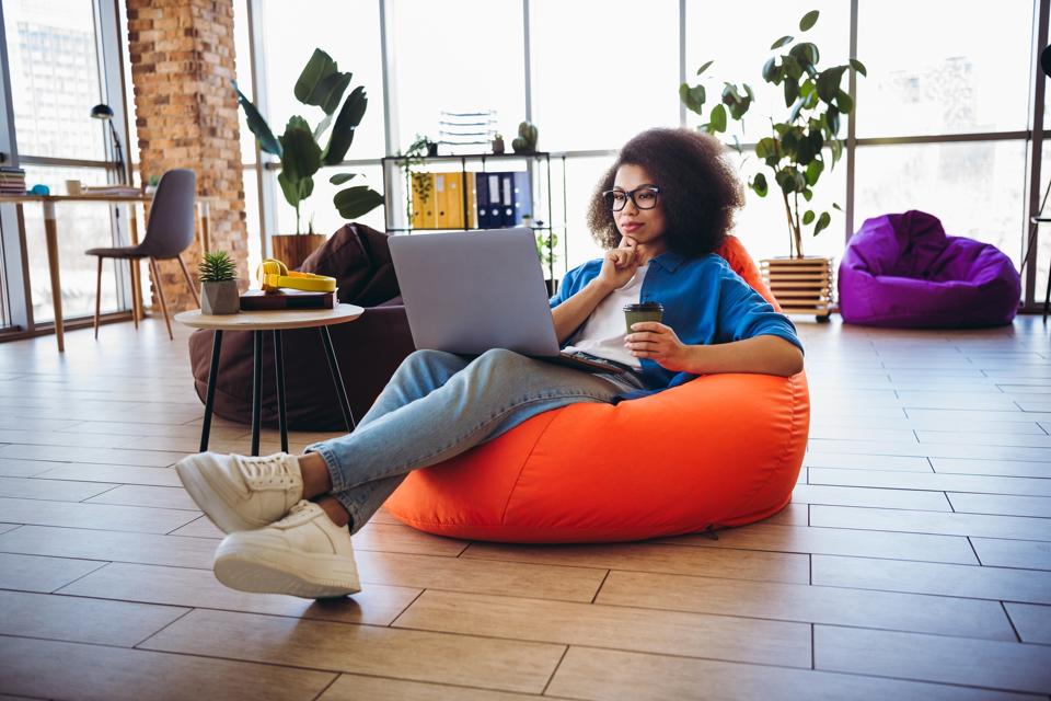 Young female professional working in a modern workspace with relaxed environment, holding a drink and using a laptop