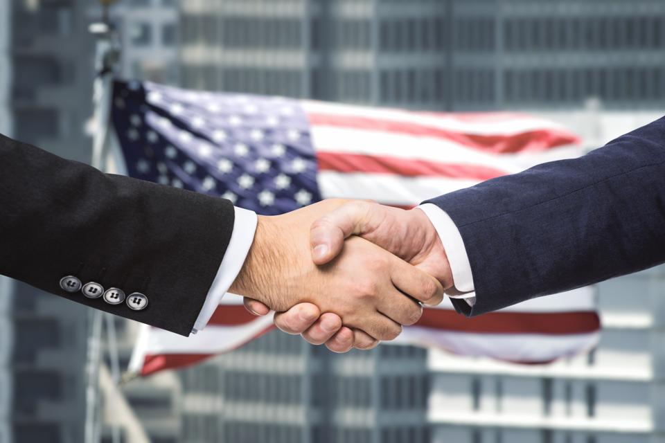 Close-up photo of leaders shaking hands after trade deal with U.S. flag in background