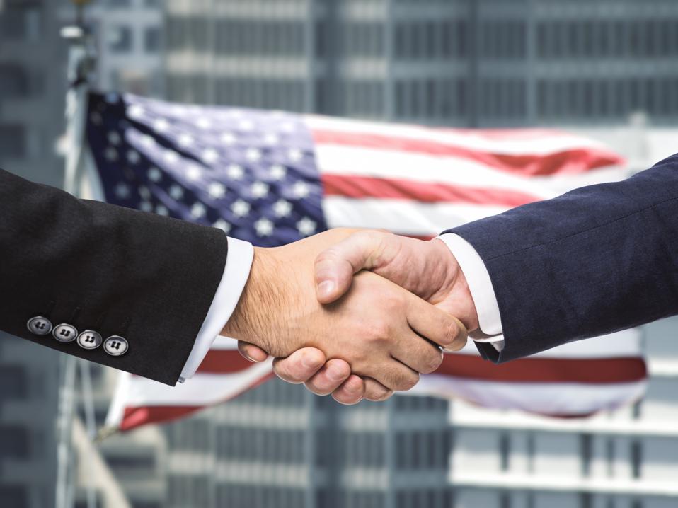 Close-up photo of leaders shaking hands after trade deal with U.S. flag in background