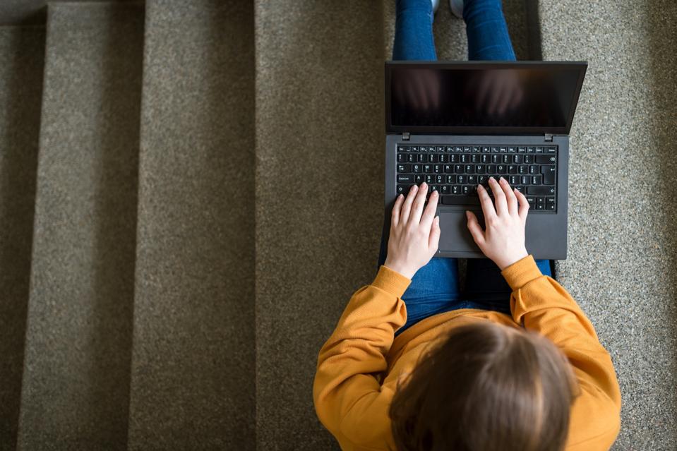 Young female college student sitting on stairs at school, writing essay on her laptop. Education concept. View from above.