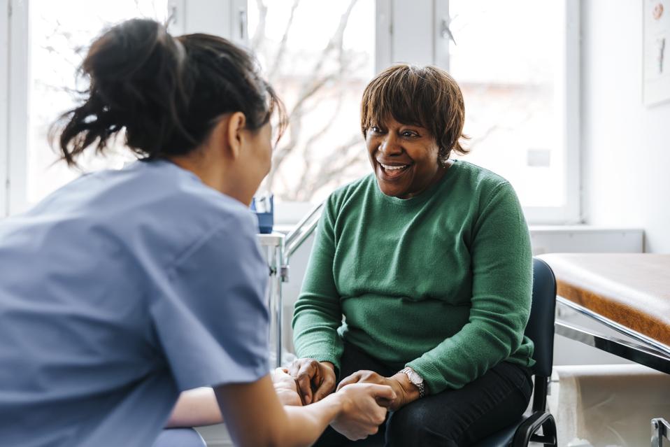 Female healthcare worker holding hands of happy senior woman sitting in examination room at hospital