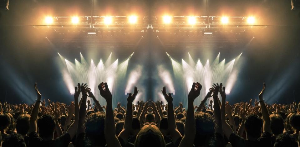 Concert stage, people are visible waving and clapping, silhouettes are visible