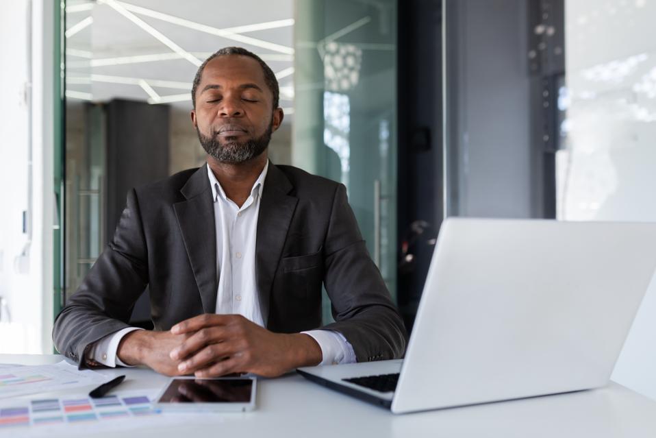 Balanced and calm man at workplace inside office, businessman meditating with closed eyes thinking and visualizing future projects and plans financial strategies, boss inside office with laptop