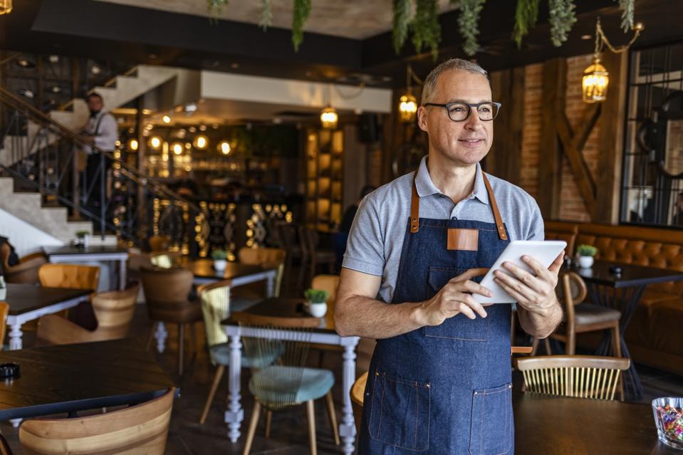 Male barista holds digital tablet in restaurant dining room.