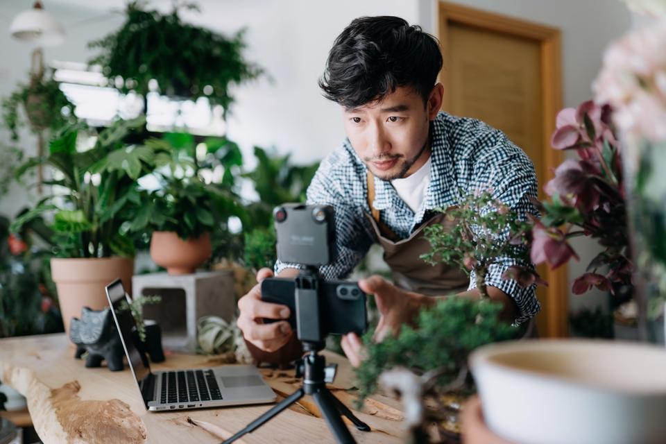 Young Asian male botanist, owner of small business flower shop, setting up smartphone lighting before starting online tutorial on potted plants arrangement, vlogging on smartphone and laptop