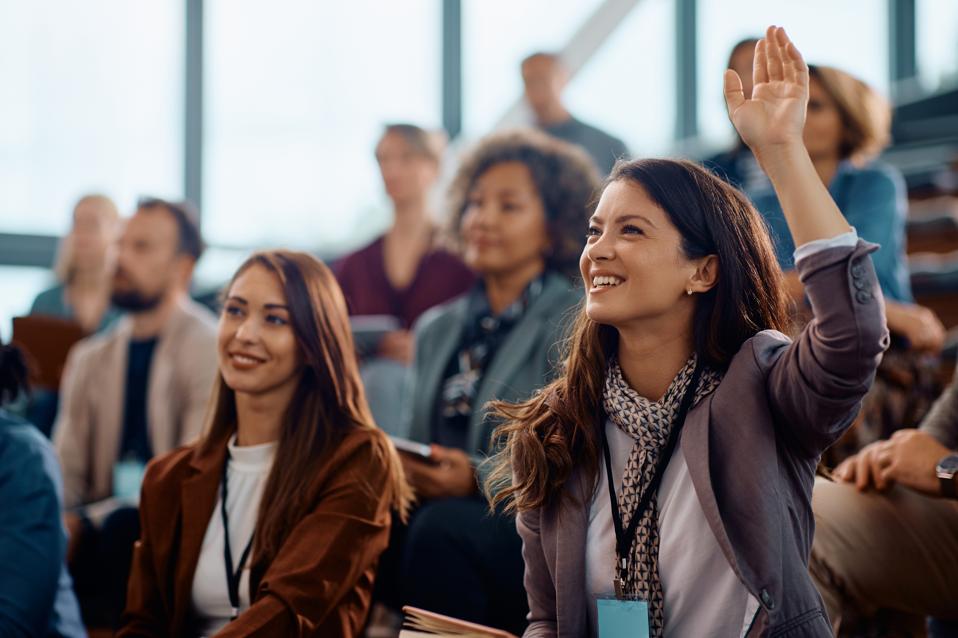 Happy entrepreneur raising her hand to answer a question during business conference in convention center.