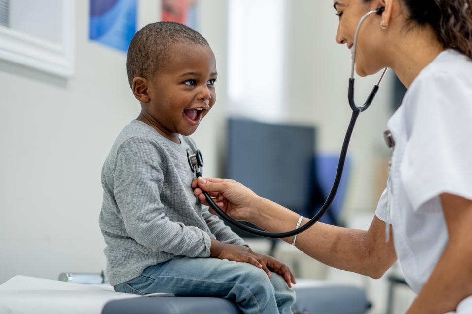 A young boy sits up on an exam table as a female Paediatrician preforms a check-up on him. 