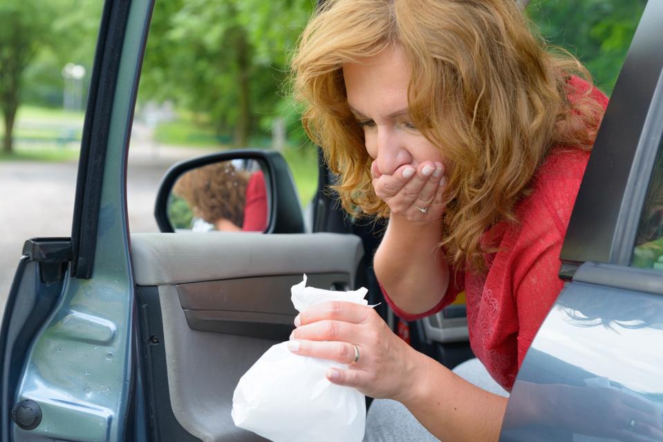 Woman leaning out of open car door, covering her mouth and holding a plastic bag.