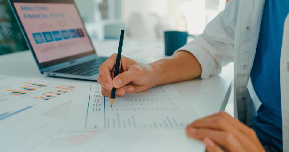 Closeup of young Asian man working from home, writing notes and analyzing financial data, showcasing remote work and financial planning. Remotely and finance.