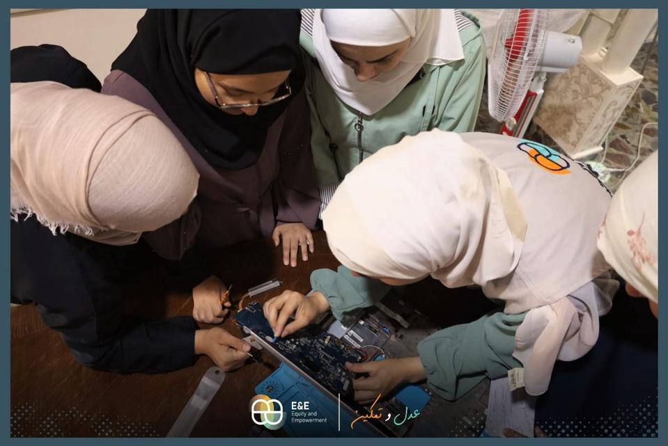 Four women wearing headscarves bend their heads over a motherboard