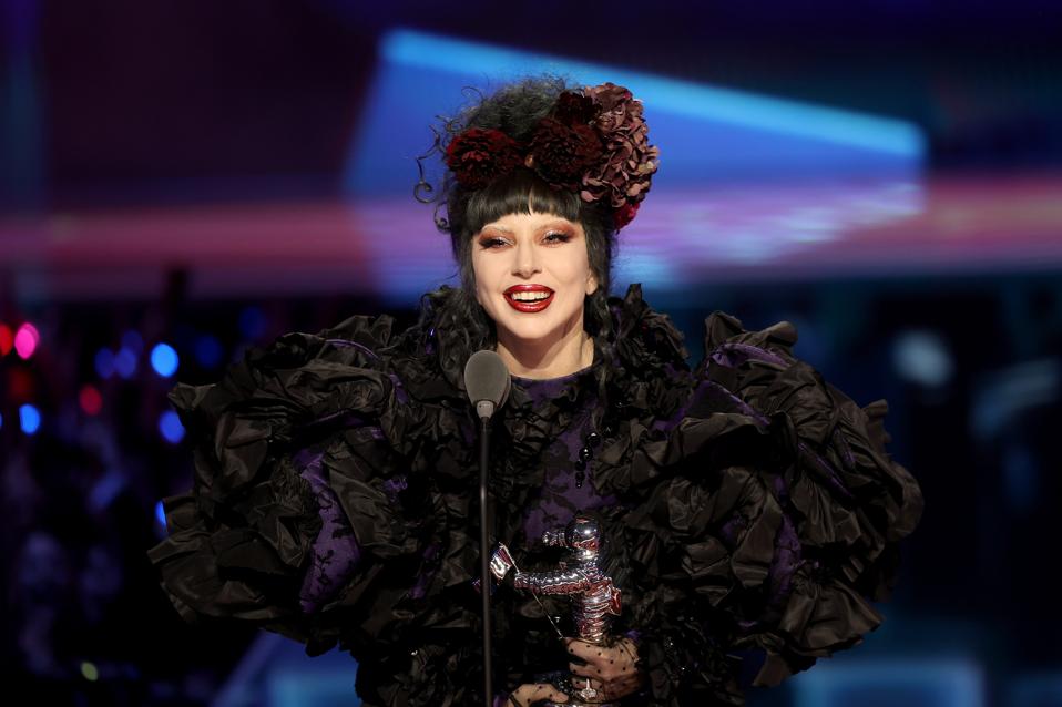 A woman with black hair and a floral headpiece holding a trophy and standing in front of a microphone