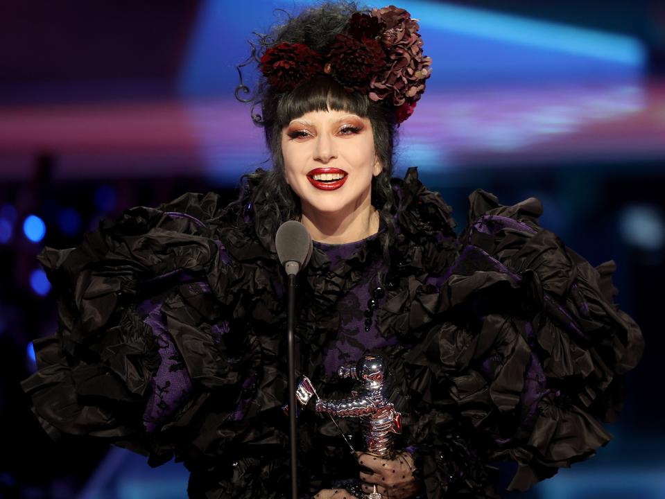 A woman with black hair and a floral headpiece holding a trophy and standing in front of a microphone