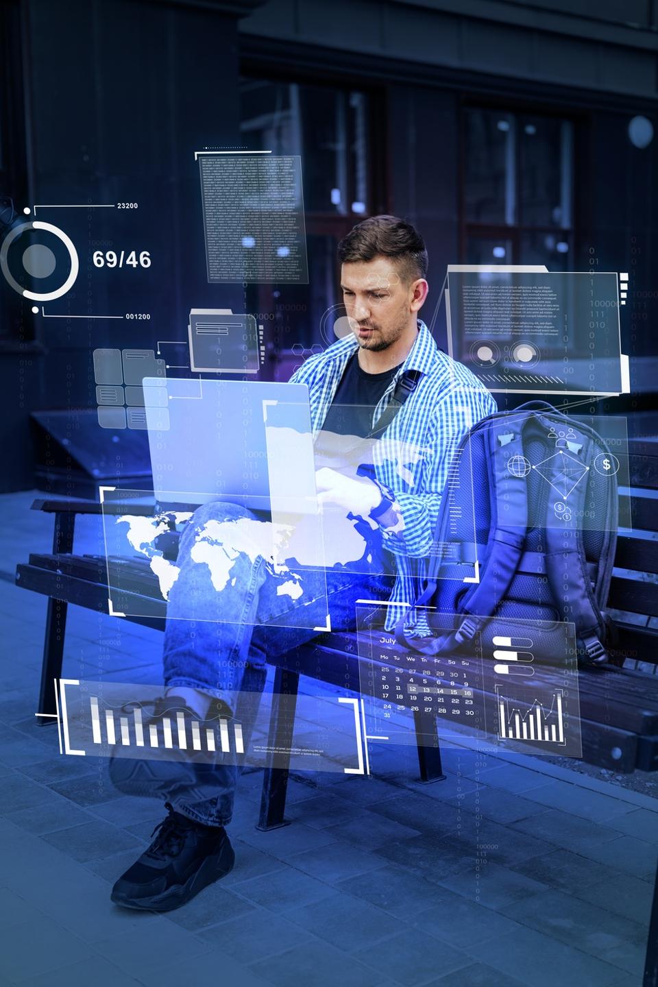 Young man using laptop in urban setting with digital interface projections during daytime