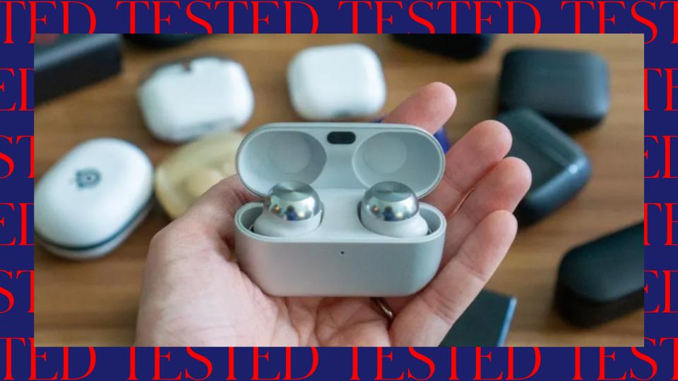 Our tech editor is holding wireless earbuds above a table full of other wireless earbuds.