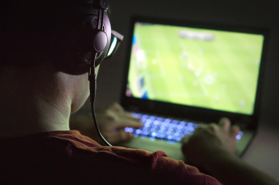Playing video games with laptop. Young man plays online soccer or football on computer. Back view of gamer with headphones in dark or late at night.