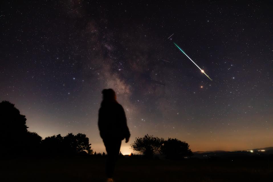 Blurry long exposure silhouette of a person looking at the Milky Way stars with meteor shower trails and countryside night landscape.