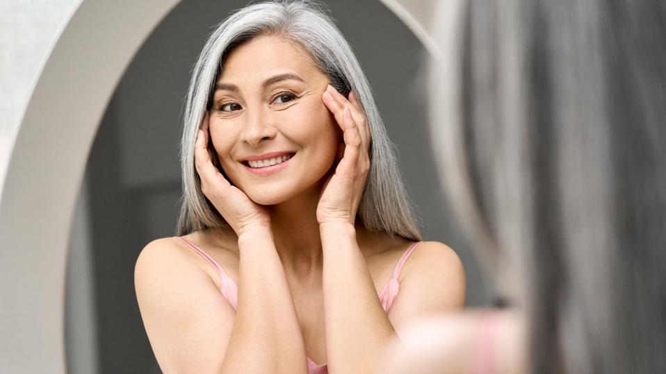 Happy mid aged Asian woman looking at mirror. Antiaging beauty care concept.