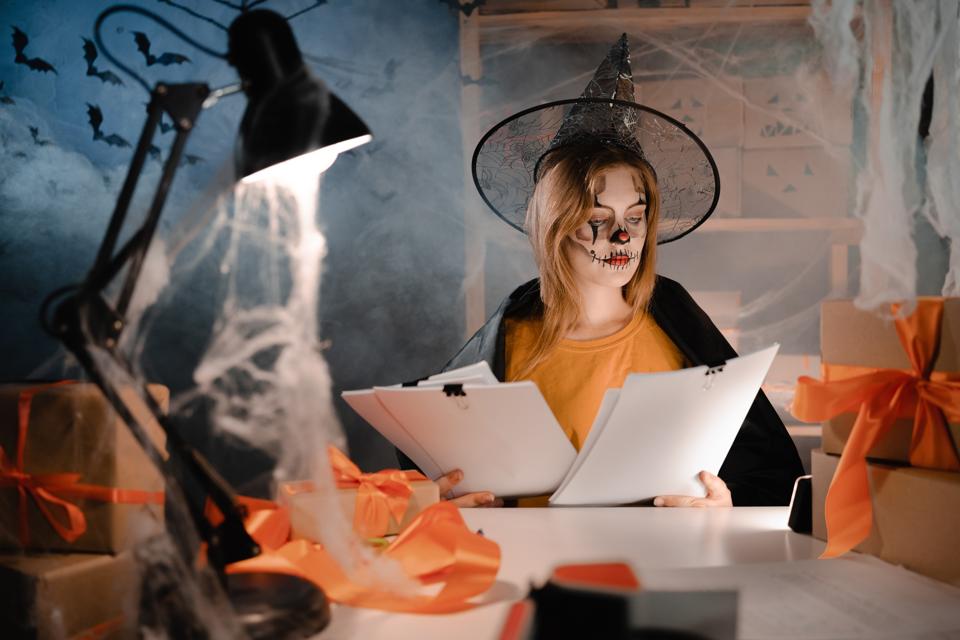 Dropshipping owner in Halloween makeup and witch costume looking at holiday sale paper report. Entrepreneur woman working at home office. Online marketing. Banner.