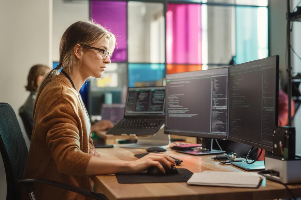 Caucasian Woman Coding on Desktop PC and Laptop Setup With Multiple Displays in Spacious Office. Female Junior Software Engineer Working on New Sprint of Mobile Application Development For Start-up.
