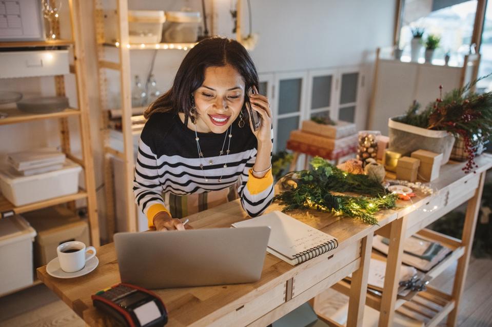 Small business owner talks on phone and checks her holiday marketing plans on her laptop.
