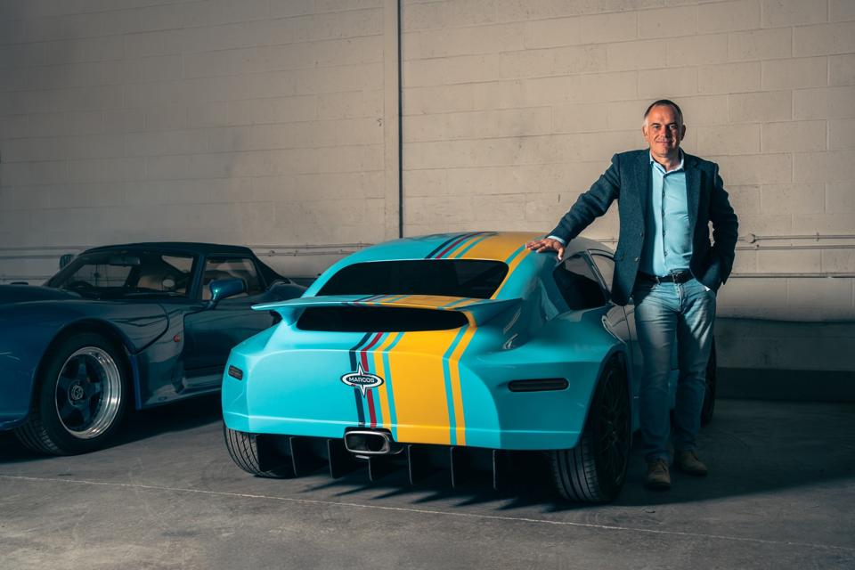 A man with jeans and a jacket on stood next to the rear of a blue and yellow car 