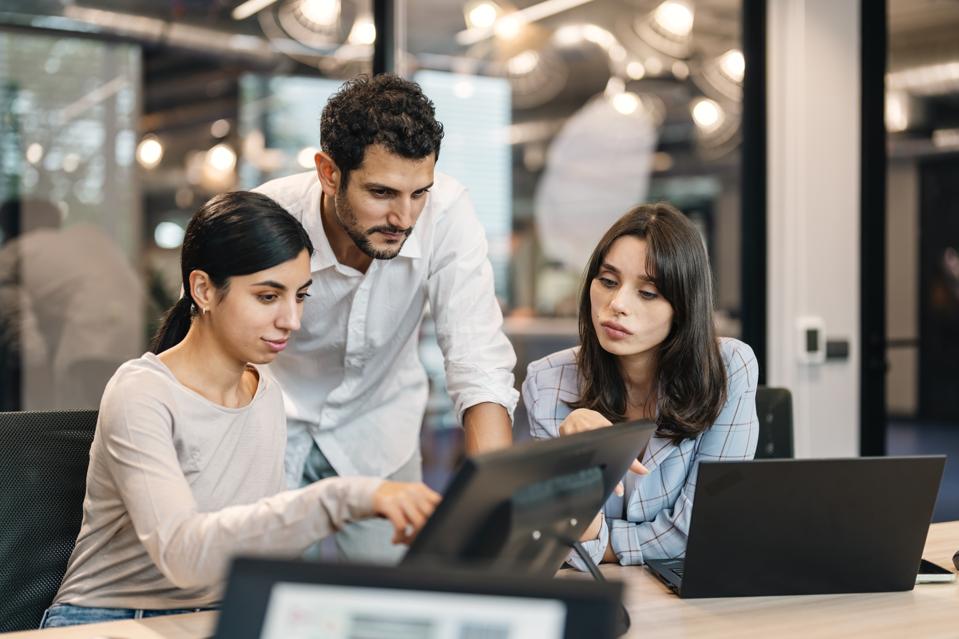 Group of business people working, the team are sitting at a board room table looking at a laptop computer. All dressed in casual business clothing with a window behind them.