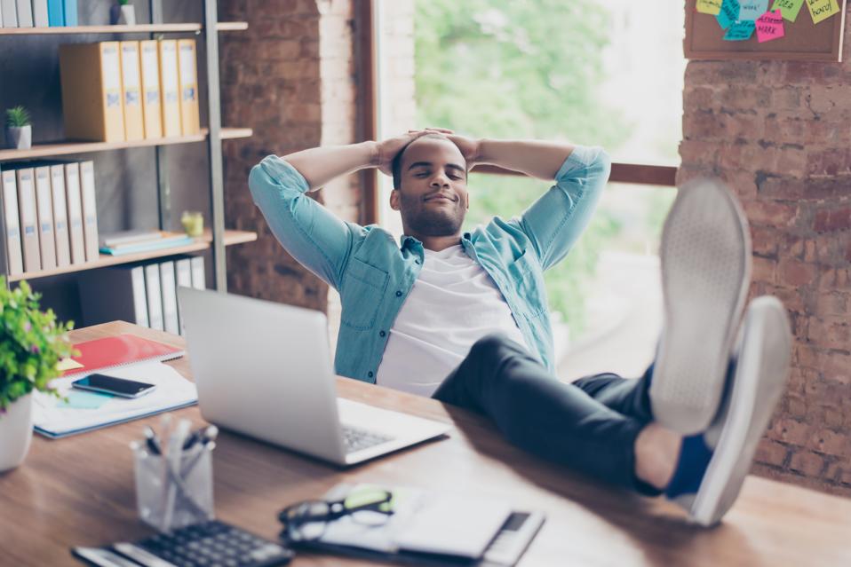 Professional Black man in light blue shirt taking a restorative break at his modern office, leaning back in his chair with hands behind his head and eyes closed, feet up on desk. The workspace features warm lighting, exposed brick walls, bookshelves, and natural elements, demonstrating a neuro-inclusive office environment that supports different working styles and the importance of mental breaks during the workday