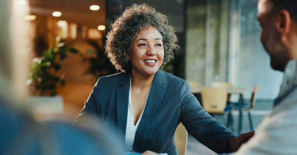 A woman in business professional smiles across the table in a meeting.