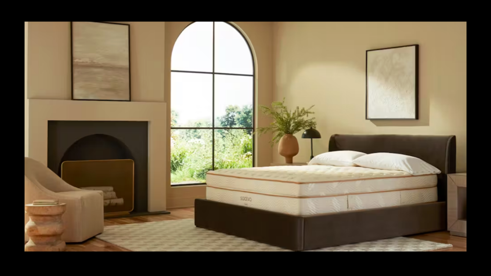 A Saatva Classic Mattress on a brown bed frame in a bedroom with a fireplace and accent chair. 