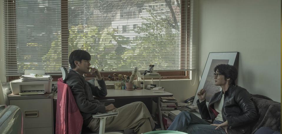 Two men talk before a wide window.