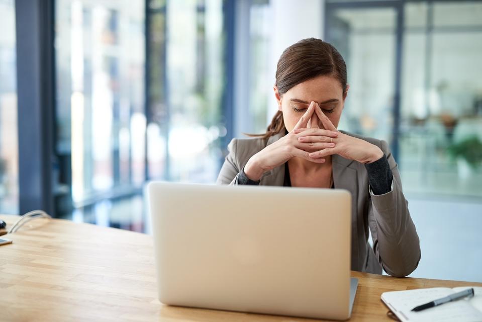 A stressed woman sits at a desk with laptop, overwhelmed by email phrases from co-workers.