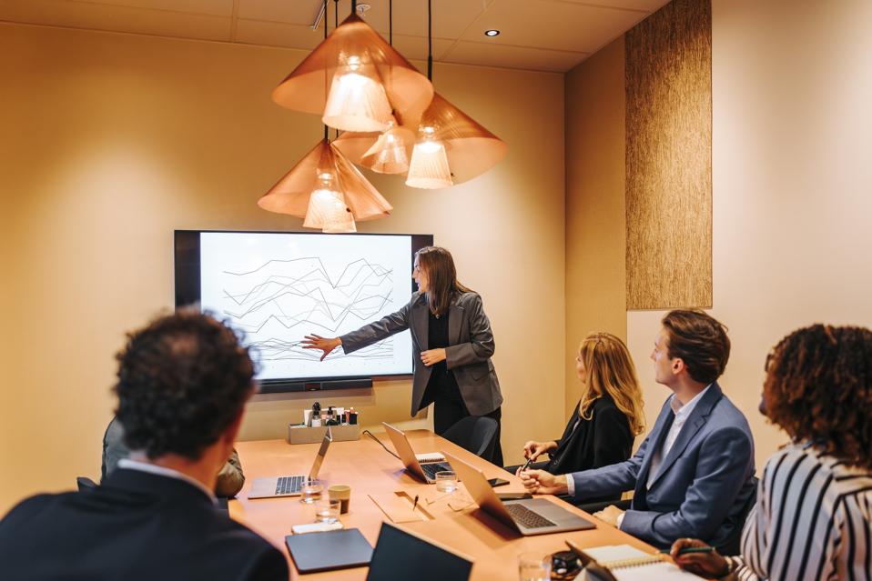 Female entrepreneur explaining graph diagram to colleagues on projection screen in board meeting at office