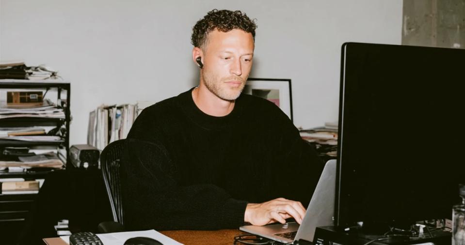 A man in a black sweater using a laptop in an office cluttered with papers