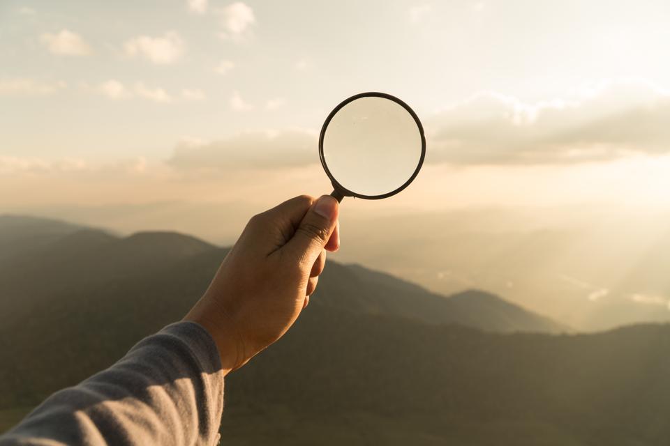 Close-Up Of Hand Holding Magnifying Glass Over Landscape Against Sky During Sunset